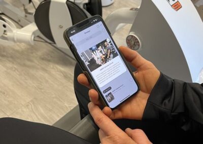 På bilden syns en person som håller i en smartphone med Endorfins träningsapp öppen. Personen sitter på en träningsmaskin, vilket antyder att appen används i samband med träning. Appens gränssnitt visar en nyhetsflödesliknande sida där användare kan interagera med innehåll relaterat till Endorfins tjänster.