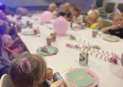 En grupp barn sitter runt ett festligt dekorerat bord med rosa ballonger och pappersdekorationer. Fokus ligger på en liten flicka med en festlig hårdekoration som dricker ur ett pappersmugg, medan de andra barnen suddigt ses i bakgrunden.