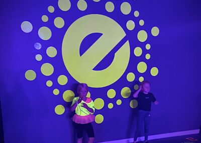 En liten flicka i en gul tröja och rosa kjol står stolt framför en stor neonljus logo på väggen i ett lekfullt gymområde, med en annan barn som tittar på intill.