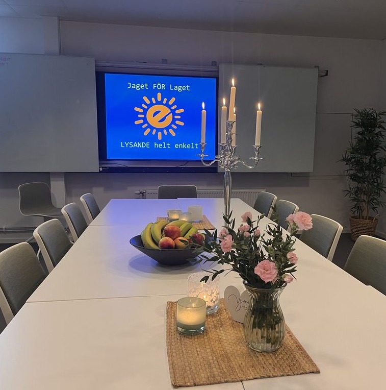 Konferensrum hos Endorfin, beläget i både Alingsås och Lerum, inrett för ett möte eller en konferens. Ett stort vitt konferensbord står i centrum, dukat med en skål fylld med färsk frukt och ett arrangemang av rosa blommor i en glasvas. På bordet finns också ett tänt ljus som bidrar till en trivsam atmosfär. I bakgrunden syns en stor skärm med Endorfins logotyp och texten 'Jaget FÖR Laget - LYSANDE helt enkelt'. Rummet framstår som välkomnande och professionellt, redo för företagsevent och samarbeten.
