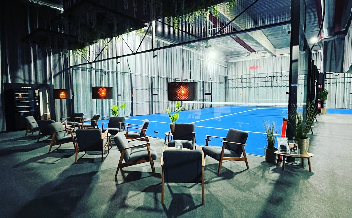Bild på en avkopplings- och loungeyta hos Padel Endorfin, en träningsanläggning belägen i Alingsås. Utrymmet är modernt och välkomnande, med eleganta trästolar och soffbord placerade på en grå matta. I bakgrunden syns en inomhus padelbana med blått golv och omgiven av nät, upplyst av varm belysning från vägglampor. Gröna växter bidrar till en lugn atmosfär där medlemmar kan slappna av före eller efter sitt träningspass.