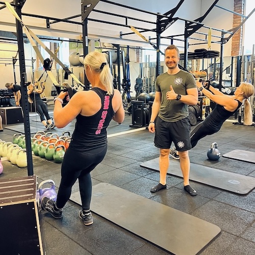 Bild på en träningsstund med personlig tränare hos Endorfin, en träningsanläggning i Alingsås och Lerum. Två kvinnor och en man utför benövningar på yogamattor. Kvinnan i förgrunden har ryggen vänd mot kameran och bär en svart träningsdräkt med texten 'Endorfin Athletics' på ryggen. Mannen i mitten ler och tummen upp, och en annan kvinna i bakgrunden utför en balansövning. I det välutrustade gymmet syns en rad kettlebells och speglar som reflekterar övriga deltagare i bakgrunden.