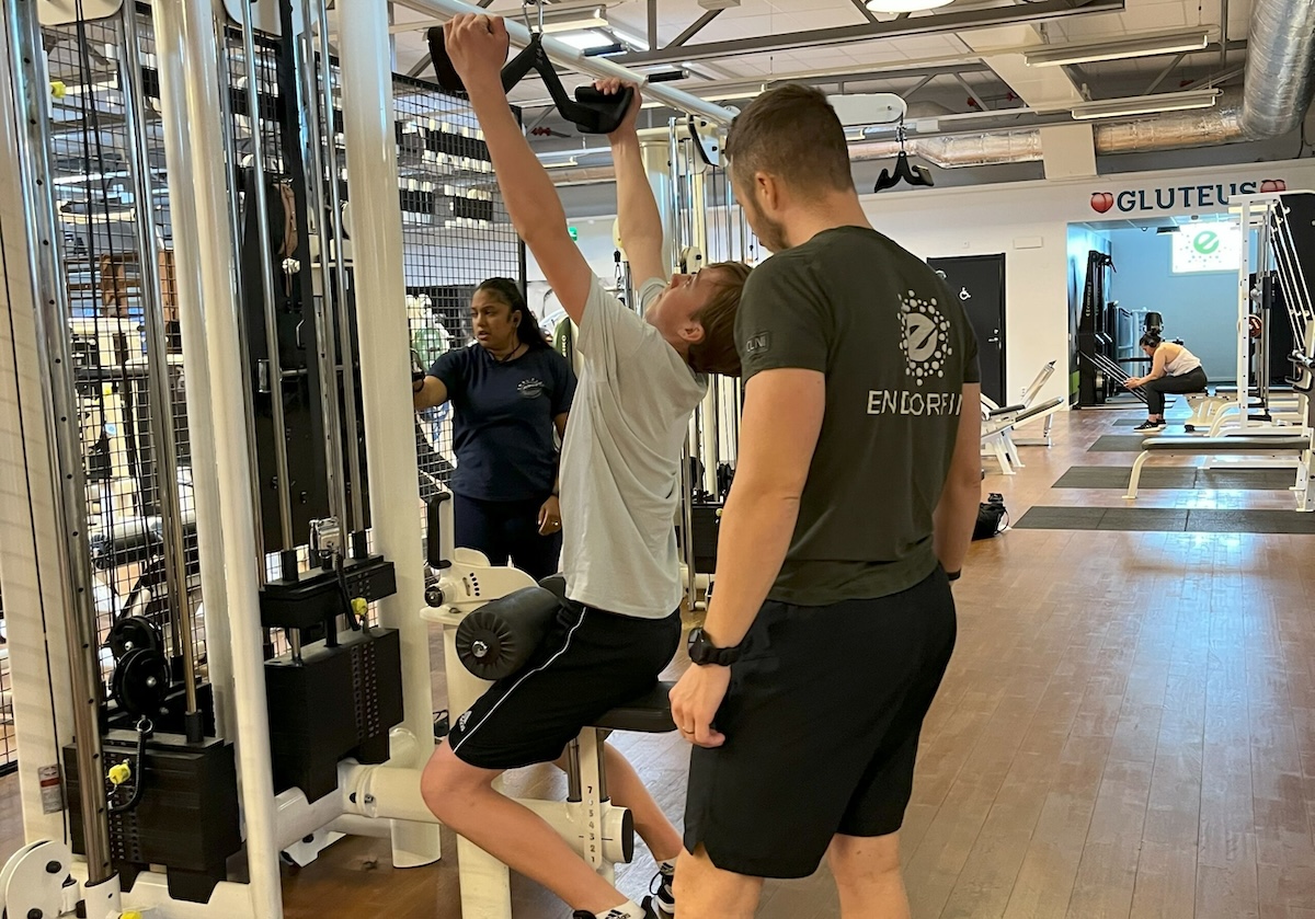 Bild från Endorfin, ett gym med verksamhet i Alingsås och Lerum, där ungdomsträning pågår. En ung deltagare, guidad av en tränare iklädd en t-shirt med Endorfins logotyp, använder ett kabelträningsmaskin för överkroppsträning. I bakgrunden observerar en annan tränare övningen. Gymmet är välutrustat och upplyst, vilket skapar en inbjudande miljö för träning och personlig utveckling för ungdomar.