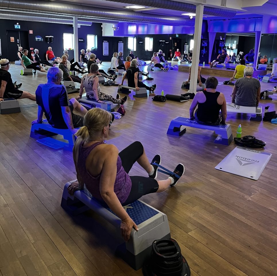 Välbesökt gruppträningsklass hos Endorfin, ett gym som finns i både Alingsås och Lerum. Deltagarna, som alla är pensionärer, sitter på step-up-brädor eller yogamattor och utför olika övningar för styrka och flexibilitet. Rummet är stort och luftigt med speglar längs väggen som skapar en känsla av rymd. Stämningen är fokuserad men avslappnad, och visar på Endorfins engagemang för att skapa en gemenskap där varje medlem kan arbeta mot sina hälsomål.